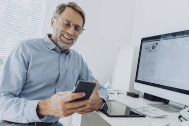 Lächelnder reifer Geschäftsmann mit Brille, der im Büro zu Hause ein Mobiltelefon benutzt - MFF07210