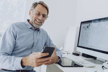 Smiling mature businessman in eyeglasses using mobile phone at home office - MFF07210