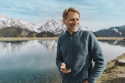 Smiling mature man holding mobile phone while standing against Prinzensee, Salzburger Land, Austria - MFF07219