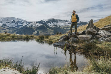 Mann mit Händen in den Taschen, stehend auf einem Felsen vor einem Teich im Salzburger Land, Leoganger Berge, Österreich - MFF07231