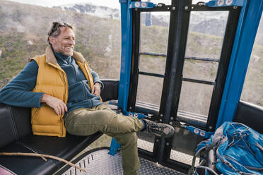 Lächelnder reifer Mann, der in einer Seilbahn sitzt und in das Salzburger Land, Österreich, blickt - MFF07246