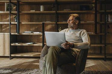 Smiling mature man with laptop sitting on armchair - MFF07247