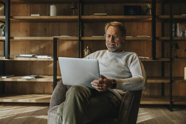 Mature man using laptop while sitting on armchair - MFF07248