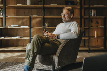 Smiling mature man using mobile phone while sitting on armchair - MFF07252