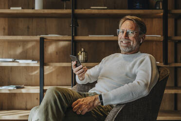 Smiling mature man holding mobile phone while sitting on armchair - MFF07253