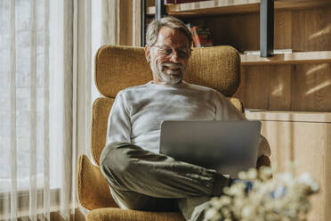 Lächelnder reifer Mann, der auf einem Stuhl sitzend am Laptop arbeitet - MFF07254