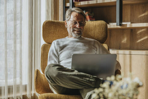 Lächelnder reifer Mann, der auf einem Stuhl sitzend am Laptop arbeitet - MFF07254