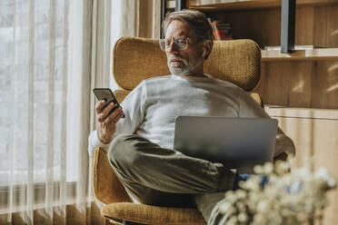 Älterer Mann mit Laptop und Mobiltelefon auf einem Stuhl sitzend - MFF07255