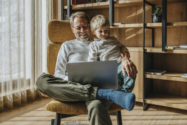Smiling mature man with son using laptop while sitting on chair - MFF07258