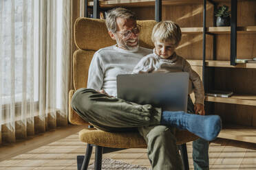 Boy looking at laptop while sitting with father on chair - MFF07259