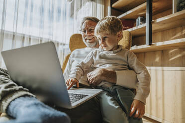 Junge benutzt Laptop, während er mit seinem Vater auf einem Stuhl sitzt - MFF07260