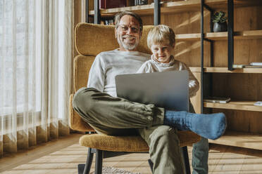 Smiling boy using laptop while sitting with father on chair - MFF07261