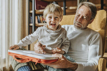 Father teaching drawing to cheerful son while sitting on chair - MFF07269