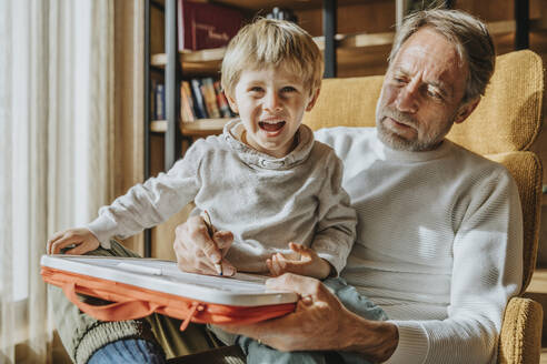 Vater bringt seinem fröhlichen Sohn auf einem Stuhl sitzend das Zeichnen bei - MFF07269