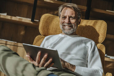 Smiling handsome man holding digital tablet while sitting on chair against shelf - MFF07272