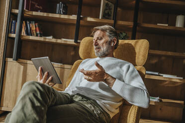 Reifer Mann gestikuliert während eines Gesprächs auf einer Videokonferenz über ein digitales Tablet zu Hause - MFF07275