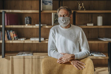 Man wearing protective face mask while standing at chair in living room - MFF07292