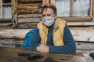 Mature man with protective face mask sitting against cottage - MFF07300