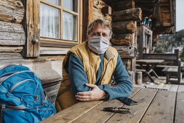 Man with protective face mask sitting by backpack against cottage - MFF07301