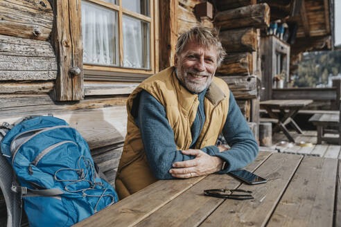 Lächelnder reifer Mann mit verschränkten Armen, der mit seinem Rucksack vor einer Hütte sitzt - MFF07302