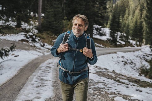 Lächelnder männlicher Tourist mit Rucksack beim Wandern auf der Straße im Winter im Salzburger Land, Österreich - MFF07307