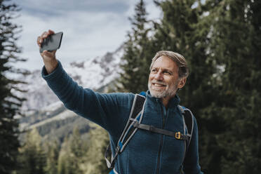 Lächelnder Mann, der mit seinem Smartphone ein Selfie vor einem Baum im Salzburger Land, Österreich, macht - MFF07309