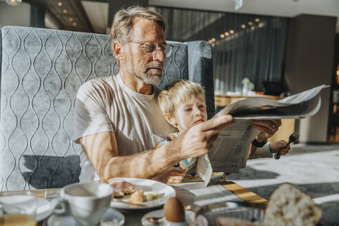Älterer Mann mit Sohn liest Zeitung, während er am Tisch im Hotelzimmer sitzt - MFF07322