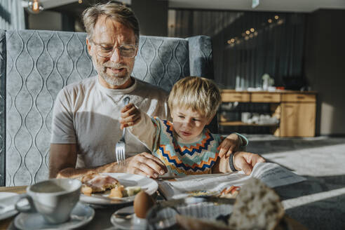 Boy eating through fork while man holding newspaper at table in hotel room - MFF07323