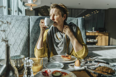 Frau trinkt Kaffee am Tisch und schaut in einem Hotelzimmer weg - MFF07327