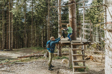 Father holding tier while son doing rope course in forest - MFF07331
