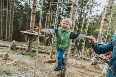 Playful boy doing rope course while supported by father in forest at Salzburger Land, Austria - MFF07337