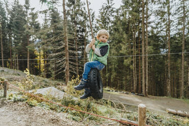 Junge sitzt auf einer Reifenschaukel im Wald während der Ferien im Salzburger Land, Österreich - MFF07339