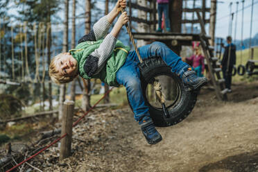 Playful boy swinging on tire swing during rope course in forest - MFF07340