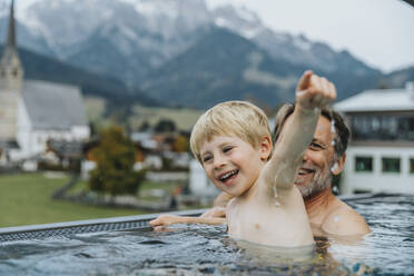 Glücklicher Vater und Sohn genießen im Infinity-Pool mit der Stadt im Hintergrund im Salzburger Land, Österreich - MFF07344