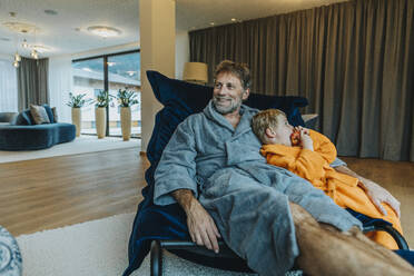 Smiling mature man in bathrobe embracing son while sitting on rocking chair in hotel room - MFF07353