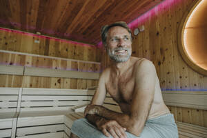 Lächelnder reifer Mann, der wegschaut, während er in der finnischen Sauna sitzt - MFF07361