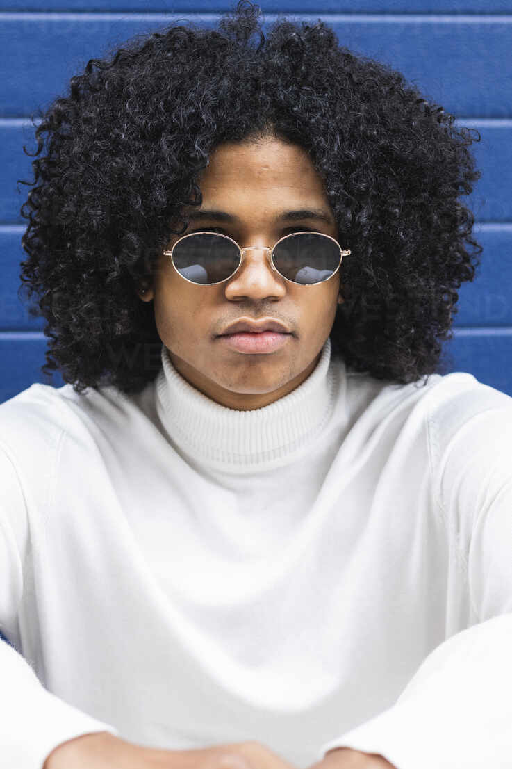 Curly hair teenage boy wearing sunglasses sitting against blue