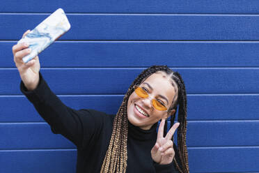 Lächelnde Frau, die ein Selfie mit ihrem Mobiltelefon macht, während sie an einer blauen Wand steht - PNAF00725