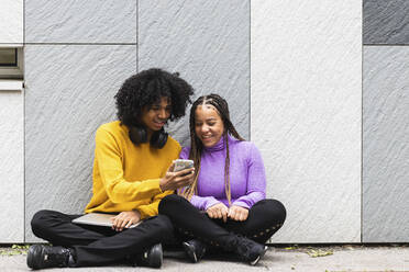 Freund zeigt seiner Freundin das Mobiltelefon, während er an der Wand sitzt - PNAF00728