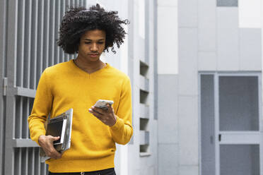 Junge mit lockigem Haar und Büchern, der sein Handy benutzt, während er in der Universität steht - PNAF00731