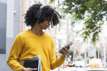 Junge mit Kopfhörern, der ein Mobiltelefon benutzt, während er mit Büchern im Freien steht - PNAF00732