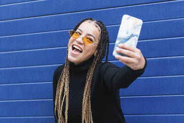 Glückliche Frau, die ein Selfie mit ihrem Mobiltelefon macht, während sie an einer blauen Wand steht - PNAF00740