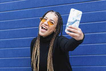 Happy woman taking selfie through mobile phone while standing against blue wall - PNAF00740