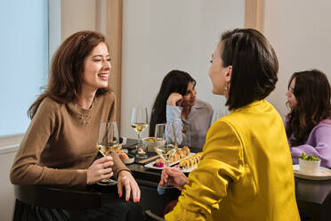 Smiling female friends with glass of white wine talking while sitting at table in restaurant - AODF00302