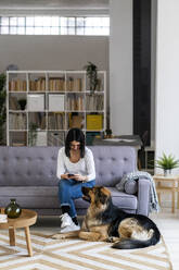 Junge Frau benutzt ihr Smartphone, während sie mit ihrem Hund im Wohnzimmer sitzt - GIOF11301