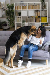 Smiling young woman playing with dog while sitting on sofa in living room - GIOF11303