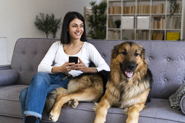 Glückliche Frau mit Smartphone, die wegschaut, während sie mit ihrem Hund zu Hause auf dem Sofa sitzt - GIOF11306