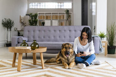 Frau, die ein Mobiltelefon benutzt, während sie mit ihrem Hund im Wohnzimmer auf dem Teppich sitzt - GIOF11308