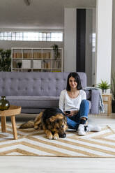 Smiling woman sitting with dog on carpet against sofa in living room - GIOF11309