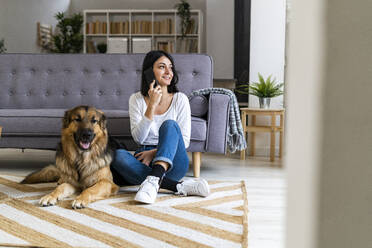 Lächelnde Frau spricht am Telefon, während sie mit ihrem Hund auf dem Teppich zu Hause sitzt - GIOF11310